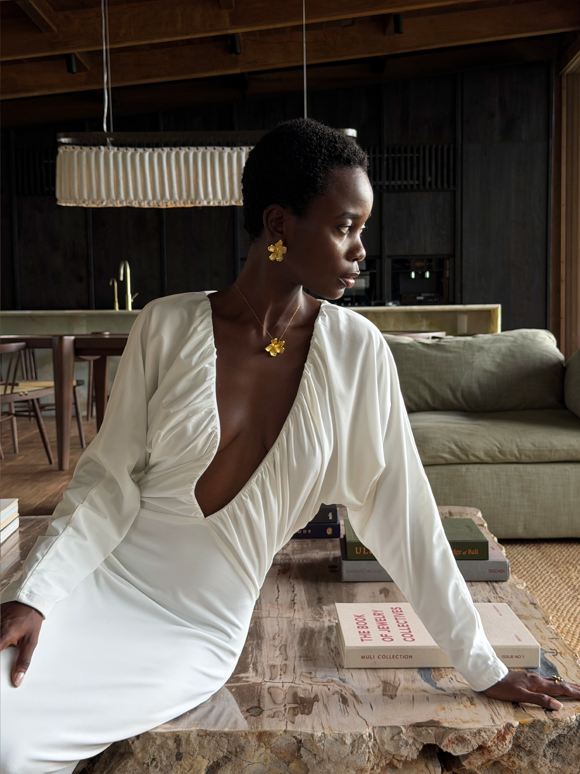 Woman in a white dress sitting at a table in a modern interior setting wearing maxi flower earrings and maxi flower necklace in gold.