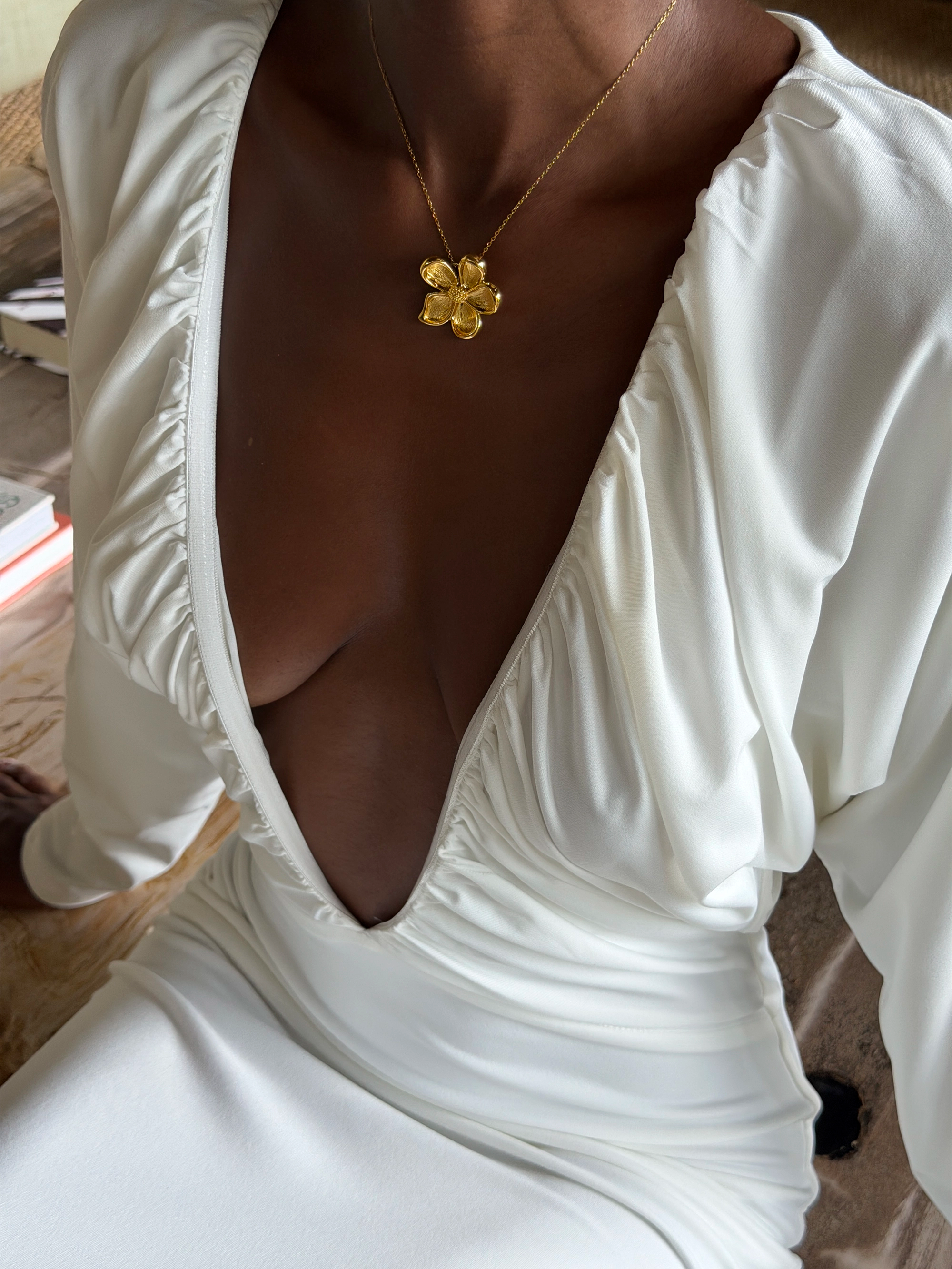 White dress with a deep neckline and gold  necklace featuring a flower pendant.