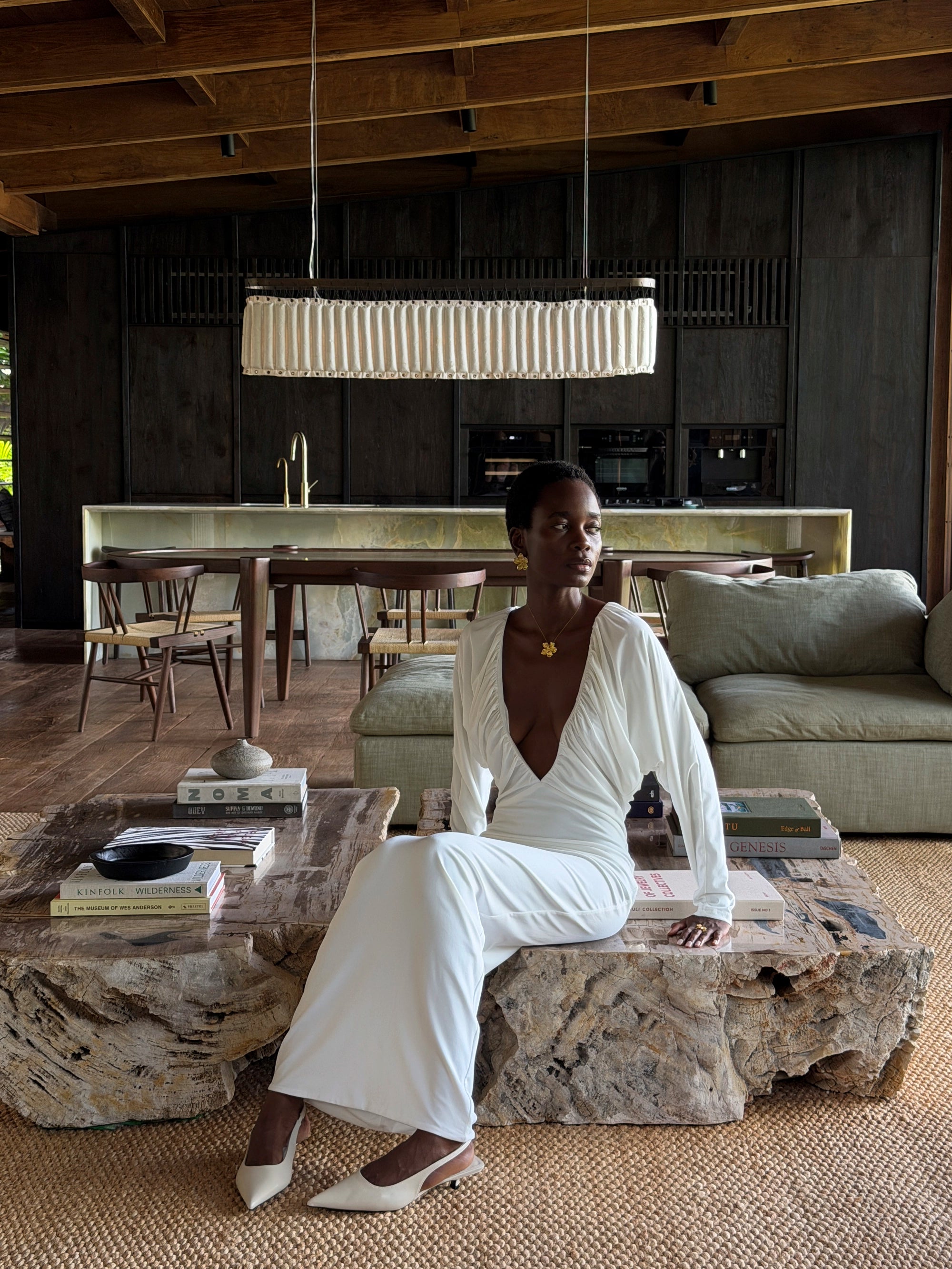 Woman in a white outfit sitting on a stone bench in a modern interior setting wearing maxi flower necklace and maxi flower earrings.